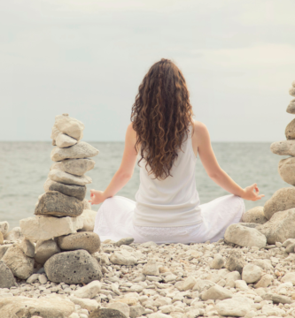 Woman meditating on a beach amongst rock cairns.