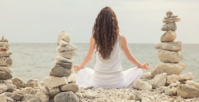 Woman meditating on a beach amongst rock cairns.